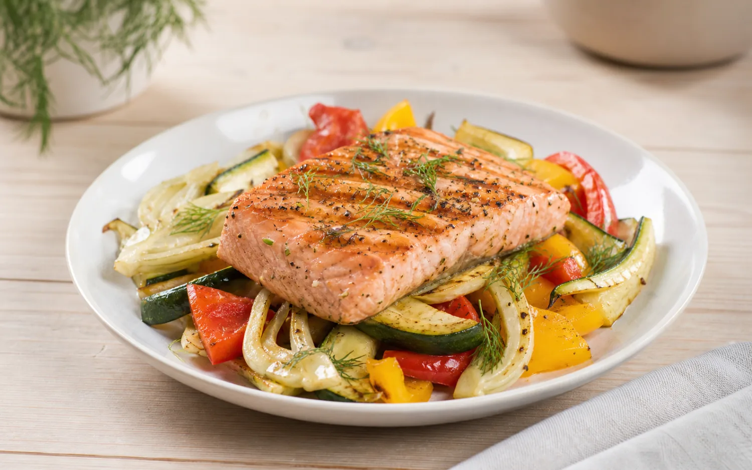 Gegrilde zalm met groenten geserveerd op wit keramisch servies op een lichte houten tafel.